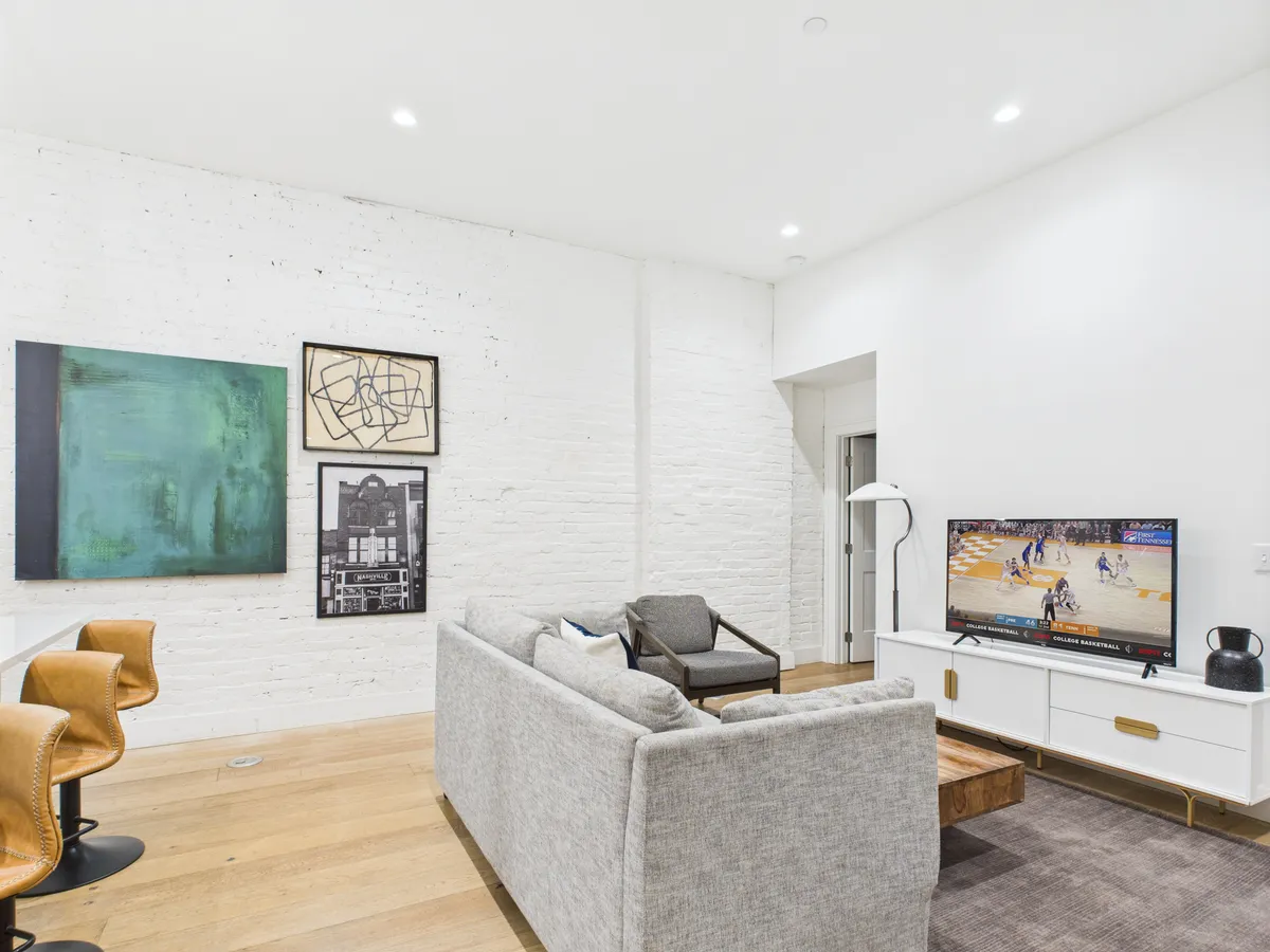 A stylish, open living area featuring a comfortable sofa, wall-mounted television, and eye-catching artwork. Bar stools and additional seating hint at a space designed for entertaining. The lighting and decor create a cozy yet lively mood, ideal for evenings spent with friends watching sports or sharing stories.