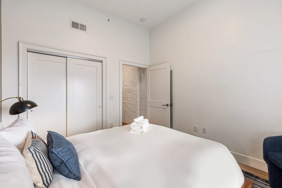 A cozy bedroom featuring a neatly made bed with white linens and decorative pillows, a desk lamp, and open closet doors showcasing ample storage.