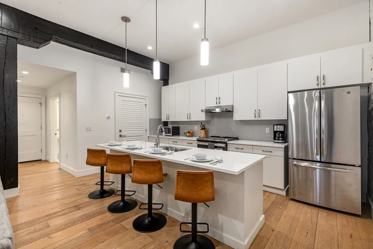 This interior scene features a welcoming, contemporary kitchen highlighted by stainless steel appliances, a central kitchen island with bar stools, and ample open counter space. The mood is lively, with elements indicating cooking, entertaining, and family gatherings, showcasing the practical and social heart of the home.
