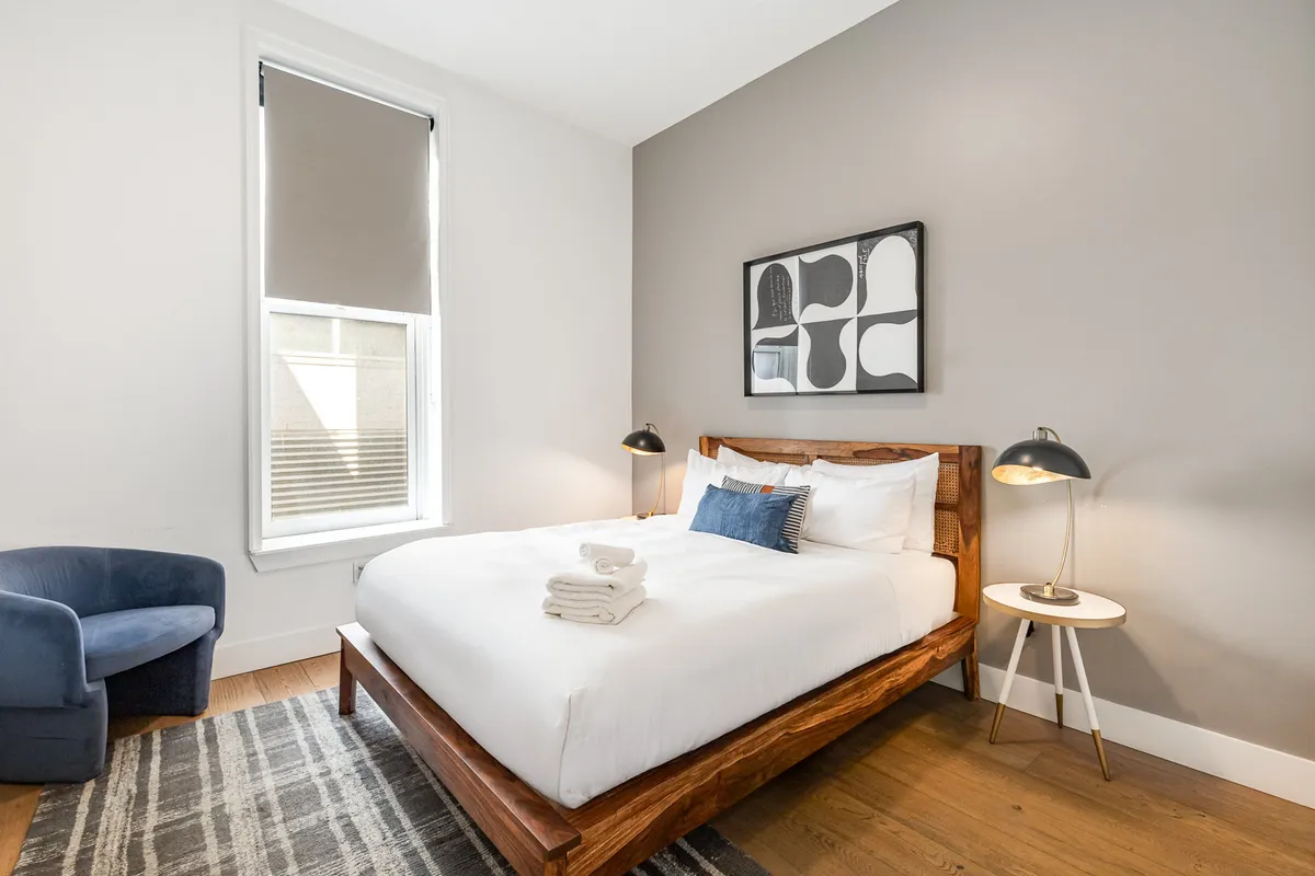 A bedroom scene with a bed dressed in crisp white linens, an abstract piece of wall art, a bedside lamp, and a blue armchair. The room receives soft natural light, accentuating the tall ceilings and bringing a sense of calm. Modern and minimalist touches are present, blending comfort with a touch of urban sophistication.