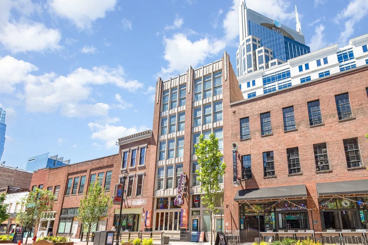An exterior view that juxtaposes the building’s historic brick façade with a modern glass skyscraper in the background. Green trees and an outdoor seating area create a lively streetscape, and the image exudes the vibrancy and walkability of the downtown neighborhood.