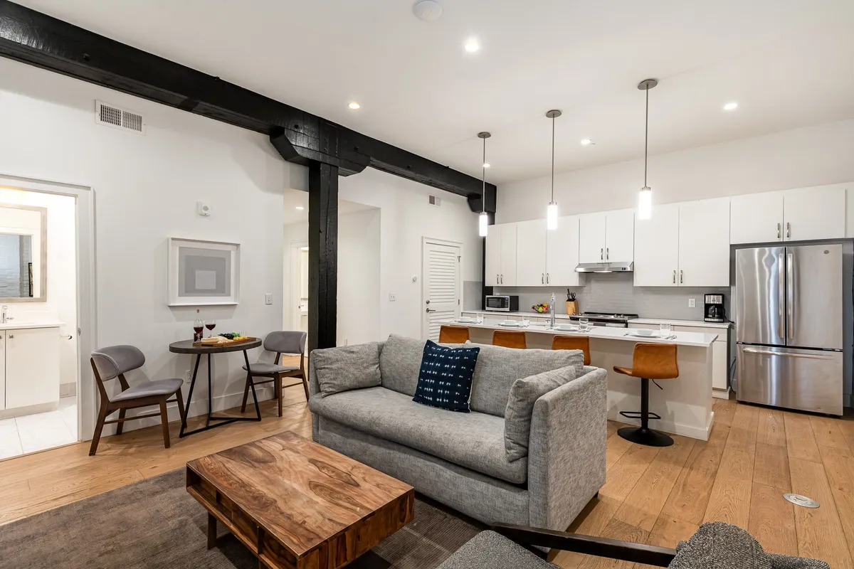 The image showcases a stylish, open-concept living space with exposed wooden beams, a plush sofa, a modern dining table, and a sleek kitchen island—all set against a backdrop of tall ceilings and warm lighting. The space invites guests to gather, dine, relax, and socialize in comfort and style.