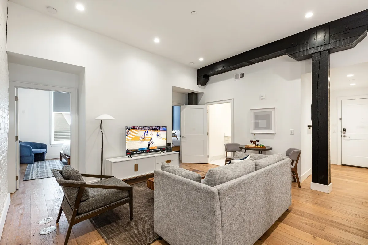 A modern living room with a comfortable sofa facing a flat-screen television. Wooden beams and a decorative lamp add character, while natural light streams in through nearby windows, highlighting the welcoming ambiance. The setup invites relaxation and shared moments, whether watching TV or enjoying each other's company.