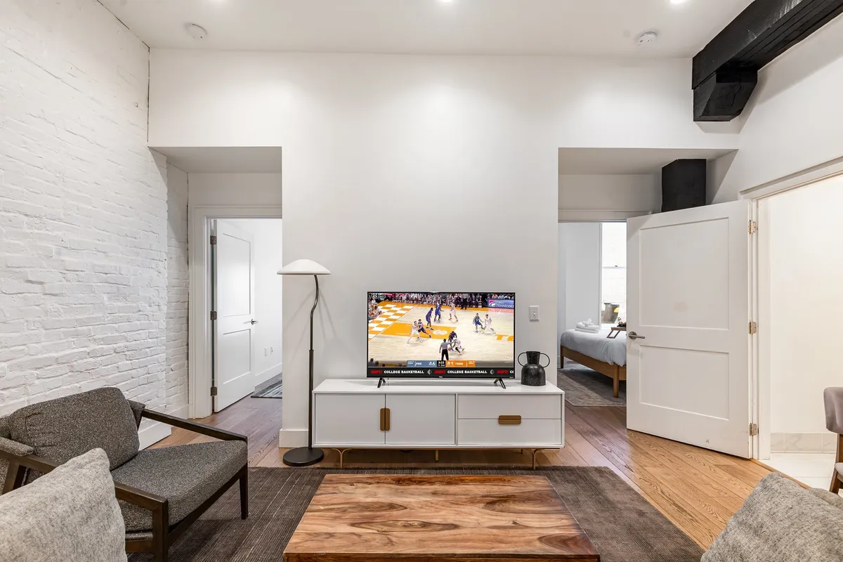 A contemporary living room with a plush sofa, modern coffee table, mounted Smart TV, and a statement lighting fixture. The room is filled with natural light and has tall ceilings, creating a spacious and airy atmosphere. The furnishings are arranged to encourage socializing, with ample seating and a casual, inviting vibe ideal for group gatherings or relaxed evenings.